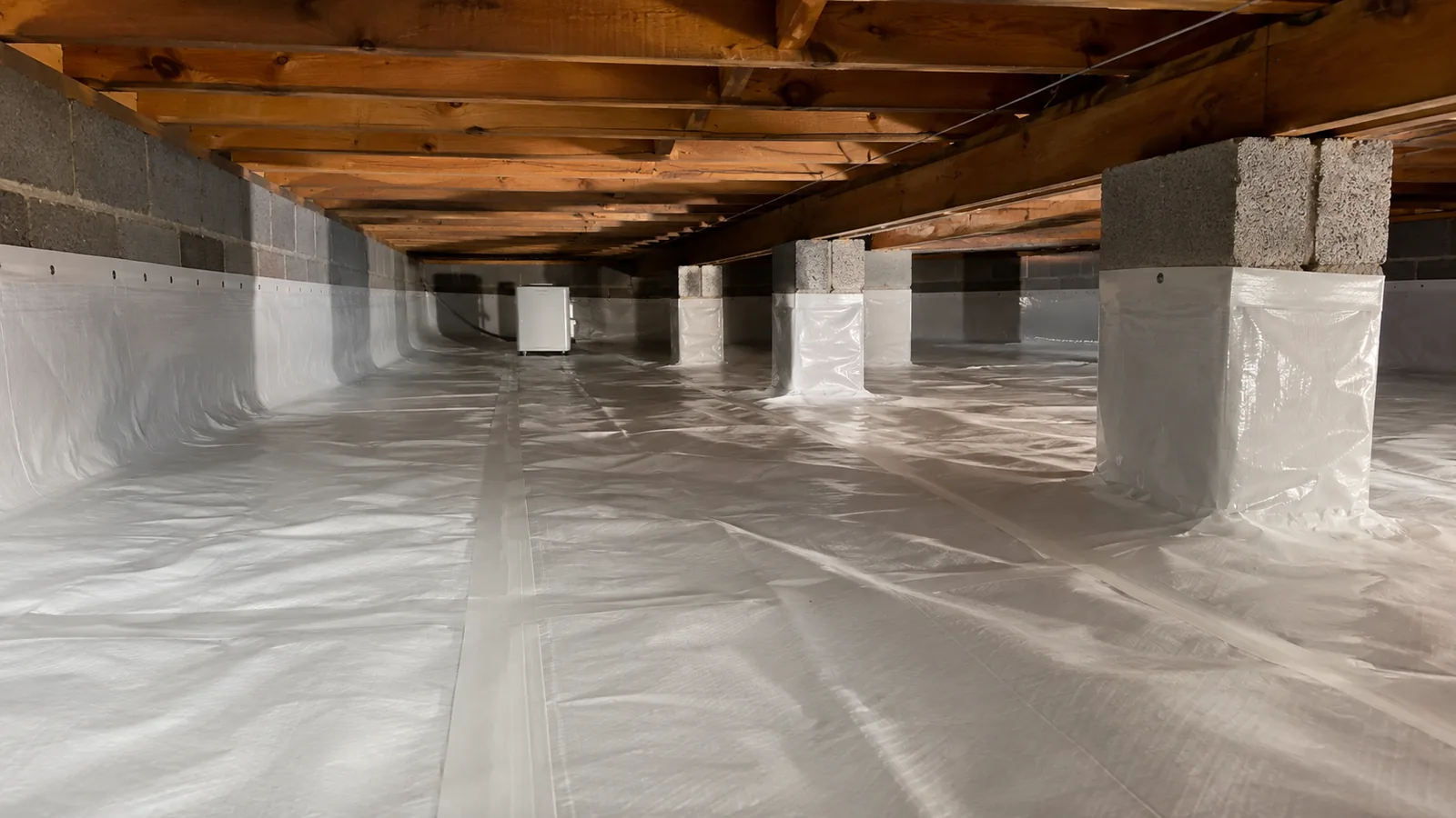 A crawl space lined with white vapor barrier across the floor and up the block piers, a dehumidifier running at the far end.