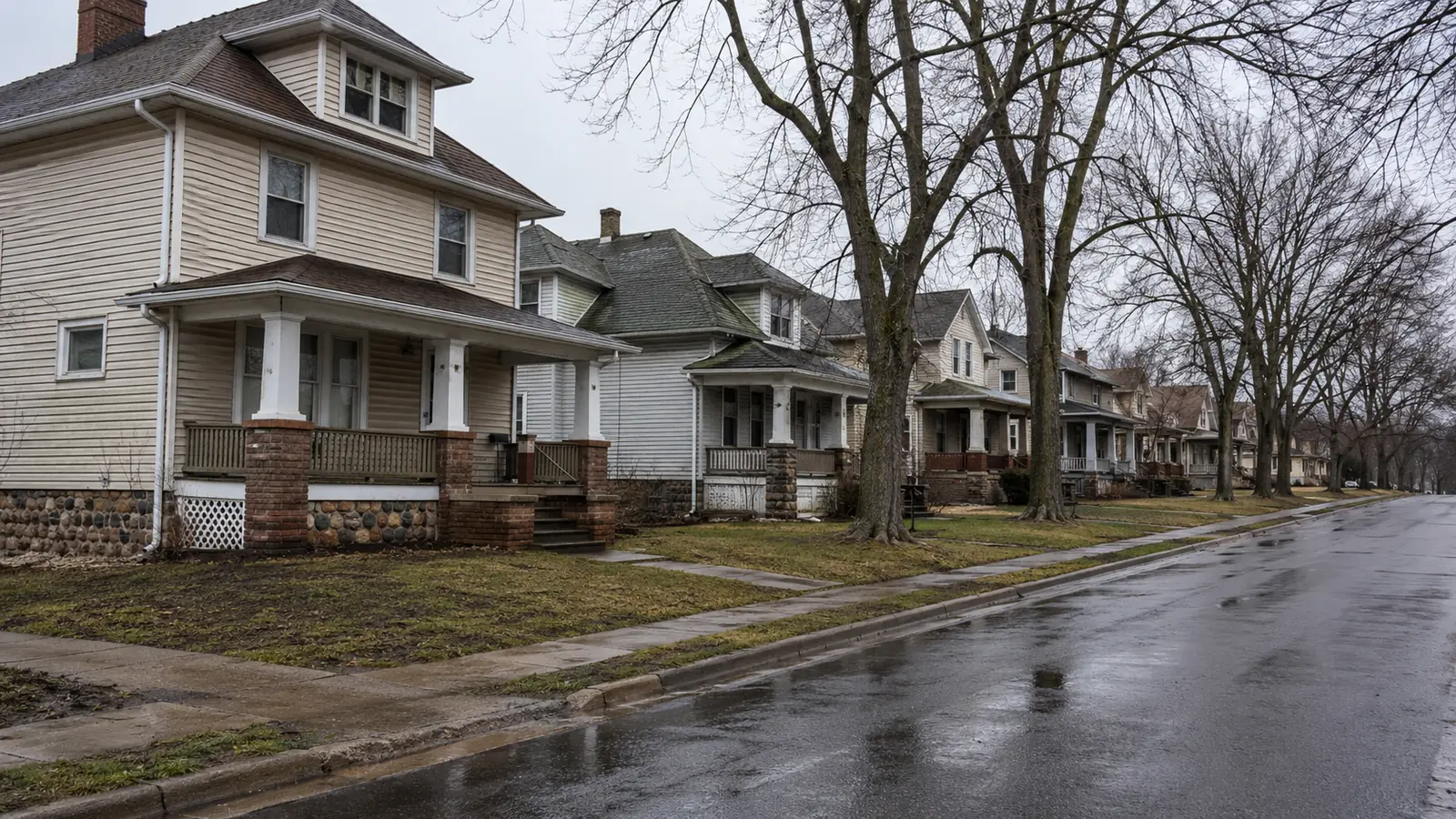 A residential street of older two-story homes with front porches on a wet overcast day.