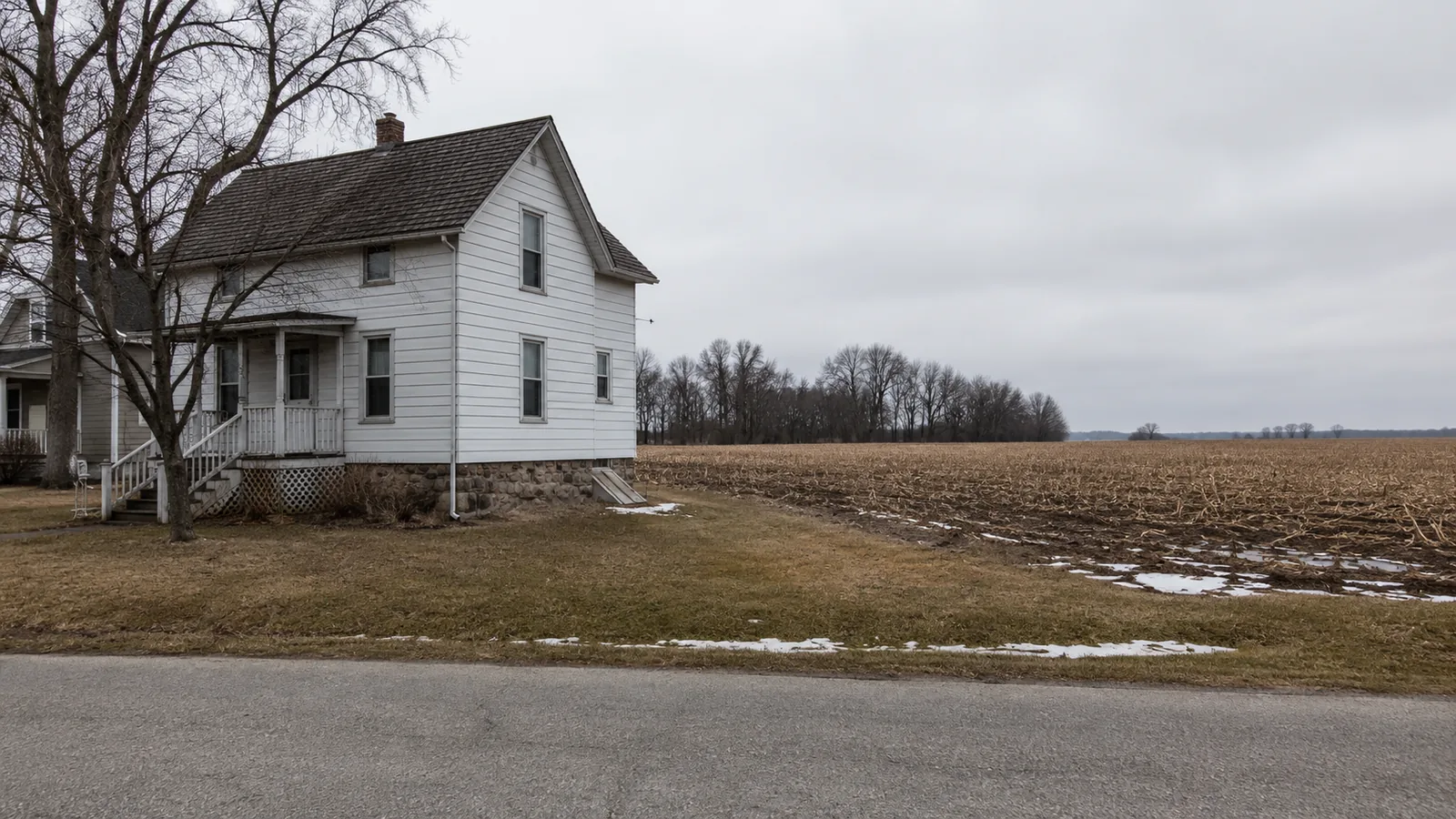 A white two-story farmhouse at the edge of a bare field on an overcast winter day.